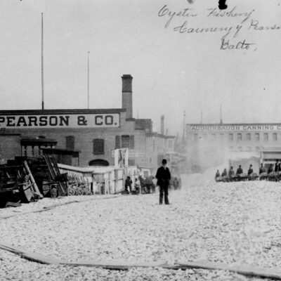 Man with a derby hat stands atop a mound of oyster shells outside the C. H. Pearson & Company oyster cannery, Baltimore. Workers bring wheel- barrows of shells from the factory to the heap. ca. 1890.